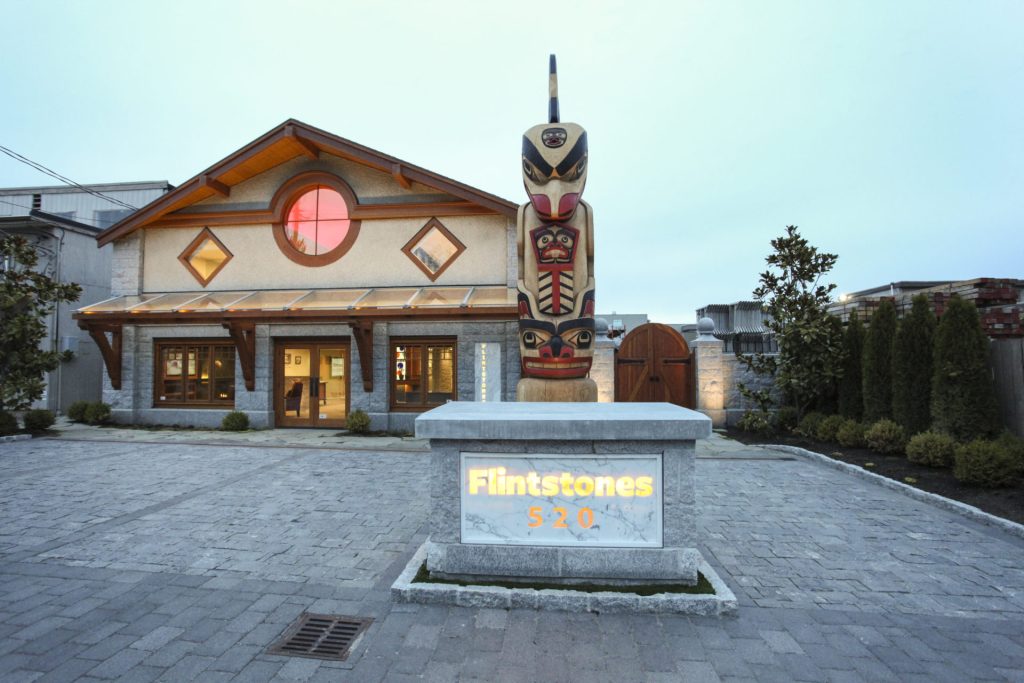 Two-level building with large stone driveway, glass awning, and totem pole